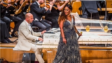 Eine Frau mit Mikrofon steht vor einem gläsernen Flügel samt Pianist auf einer Bühne 