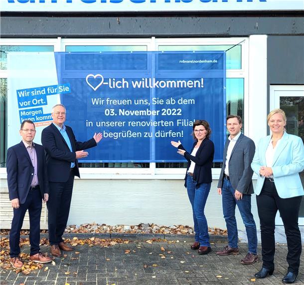 Raiffeisenbank Rodenkirchen feiert Abschluss der Renovierung Fünf Bankmitarbeiter weisen auf ein großes Plakat mit der Aufschrift "Herzlich willkommen! Wir freuen uns, Sie ab dem 3. November 2022 in unserer renovierten Filiale begrüßen zu dürfen."