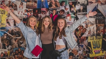Drei junge Frauen vor einer Wand mit Plakaten der 90er-Jahre.