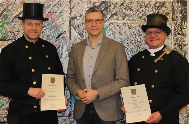 Drei Männer posieren vor einer bunt gemusterten Wand. Zwei von ihnen tragen die traditionelle schwarze Schornsteinfeger-Uniform mit Zylinder und halten ihre Bestellungsurkunden in der Hand. Der Mann in der Mitte trägt ein graues Sakko und gratuliert den beiden.