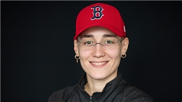 Das Gesicht einer jungen Frau, die Brille, goldene Ohrringe und eine rote Baseballkappe trägt.