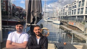 Die beiden Brüder stehen im Hafen. Im Hintergrund sind Schiffe zu sehen.