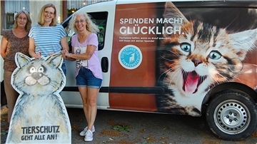 Drei Frauen stehen vor einem Transporter mit der Aufschrift „Spenden macht glücklich“ und einem großen Katzenmotiv. Sie halten ein gemaltes Pappschild mit der Aufschrift „Tierschutz geht alle an!“ in Form einer Katze.