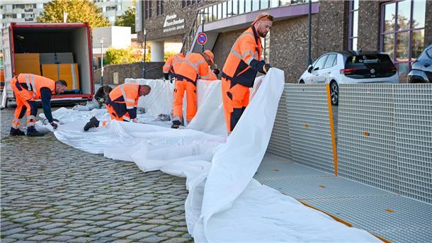Aufbau Mobildeiche  Sechs Mitarbeiter greifen sich Kunststoff-Gitter-Elemente aus einem Container, stecken sie im 90-Grad-Winkel zu einer Barriere zusammen und spannen eine Folie darüber. 
