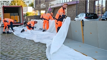 Aufbau Mobildeiche  Sechs Mitarbeiter greifen sich Kunststoff-Gitter-Elemente aus einem Container, stecken sie im 90-Grad-Winkel zu einer Barriere zusammen und spannen eine Folie darüber. 
