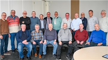 Gruppenfoto von 18 älteren Männern in Freizeitkleidung, aufgenommen in einem hellen Raum. Einige sitzen, andere stehen dahinter. 