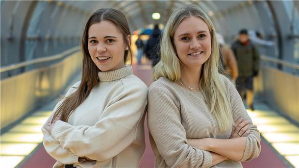 Zwei junge Frauen stehen in einem modernen, hell beleuchteten Tunnel nebeneinander und lächeln selbstbewusst in die Kamera. Beide tragen beige Pullover und haben die Arme verschränkt. 