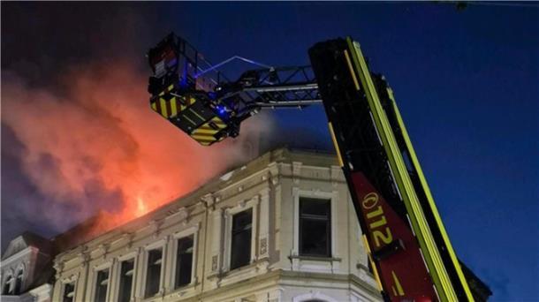 Flammen schlagen aus einem Haus im Hintergrund. Im Vordergrund ist die Drehleiter der feuerwehr zu sehen.