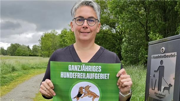 Eine Frau hält einen Zettel in der Hand, auf dem zwei spielende Hunde und der Schriftzug "Ganzjähriges Hundefreilaufgebiet" zu sehen sind. Die Stadt will ihre Freilaufflächen besser kennzeichnen.