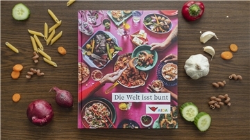 Das Kochbuch "Die Welt isst bunt" auf einem Holztisch, umgeben von Gemüse und Kochzutaten. 