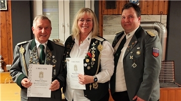 Drei Personen in traditionellen Schützenuniformen stehen in einem Raum mit Holzwänden. Sie tragen zahlreiche Abzeichen und Orden an ihren Jacken und Westen. Die beiden Personen links, ein älterer Mann und eine Frau mit blonden Haaren, halten jeweils eine Urkunde.