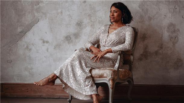 Vollmond-Lounge im Pferdestall: Malia präsentiert Neo Soul-Jazz Eine dunkelhäutige Frau in einem schimmernden Kleid sitzt auf einem Stuhl.