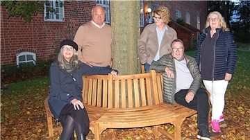Rotary Club Hagen: Spende für die Burg und Geschenke für Lkw-Fahrer Fünf Personen stehen und sitzen um eine halbrunde Holz-Baumbank im herbstlich gefärbten Burghof. Im Hintergrund ist ein historisches Backsteingebäude zu sehen.