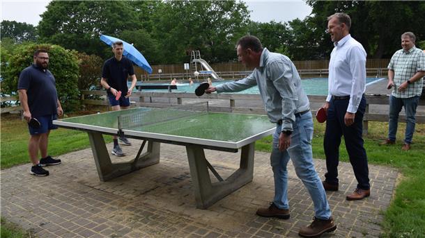 Fünf Männer spielen an einer Tischtennisplatte im Freien, im Hintergrund schwimmen Menschen in einem Freibad.