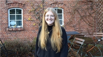 Auf dem Foto abgebildet ist die lächelnde FSJlerin Wiebke Tüchter, 20 Jahre alt, lange blonde Haare, Brille mit hellem Gestell, in einem schwarzen Pullover und goldener Halskette mit viereckigem, goldenen Anhänger, vor der Backsteinwand des Kulturzentrums Seefelder Mühle.