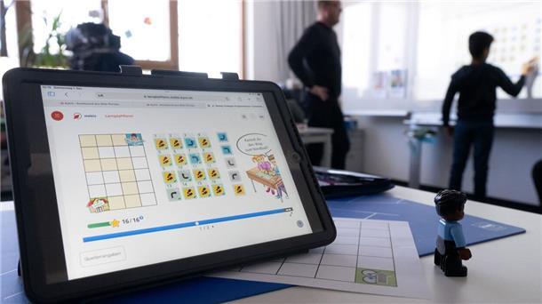 Ein Tablet in einem Klassenzimmer, im Hintergrund ein Schüler und ein Lehrer an der Tafel.
