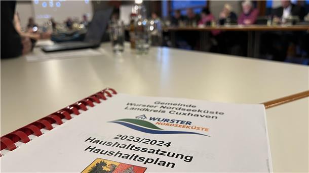 Im Vordergrund liegt der Haushaltsplan Hefter, im Hintergrund sind verschwommen Menschen an Tischen zu erkennen.