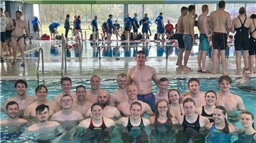 Gruppenfoto von rund 25 jungen Erwachsenen der DLRG Hagen im Bremischen, die in Badekleidung im Schwimmbecken stehen und lächeln; im Hintergrund weitere Wettkampfteilnehmende am Beckenrand einer Schwimmhalle.
