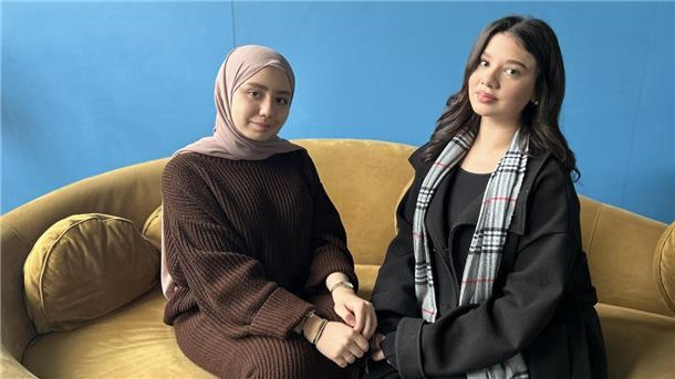 Schwestern mit syrischen Wurzeln erhalten Stipendium Zwei junge Frauen sitzen vor einer blauen Wand auf einem senfgelben Sofa. Die Frau links trägt ein braunes, langärmeliges Kleid und ein taupefarbenes Kopftuch. Die Frau rechts hat schwarze Haare, trägt einen schwarzen Mantel und einen karierten Schal.