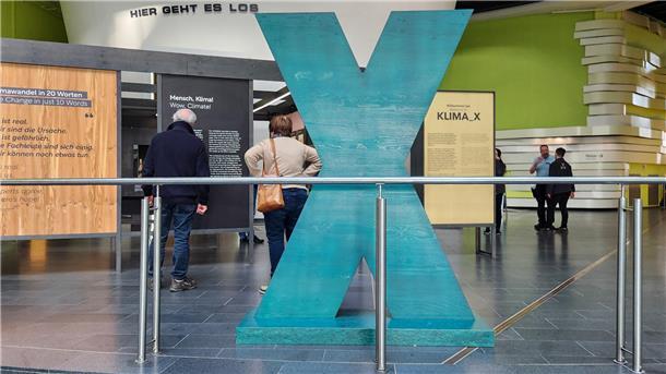 Besucher betrachten die Ausstellung „KLIMA_X“ im Klimahaus Bremerhaven. Im Vordergrund steht ein großes, türkisfarbenes X als zentrales Ausstellungselement.
