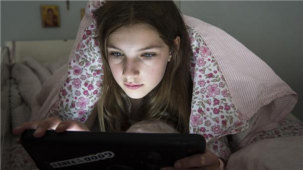 Ein Mädchen liegt auf dem Bett unter der Decke und schaut auf ein Tablet.
