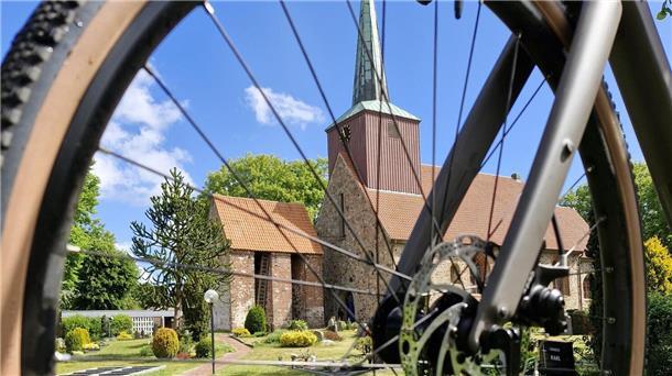 Ein Kirchengebäude mit rotem Ziegeldach und hohem Turm ist durch die Speichen eines Fahrrad-Vorderrads fotografiert. Im Vordergrund liegt ein gepflegter Friedhof.