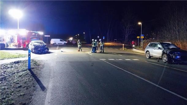 Eine nächtliche Unfallstelle an einer Kreuzung: Zwei stark beschädigte Fahrzeuge stehen auf der Straße, während Feuerwehrleute in Einsatzkleidung die Unfallstelle sichern und Betriebsstoffe aufnehmen. Im Hintergrund ist ein Feuerwehrfahrzeug mit eingeschaltetem Licht zu sehen.