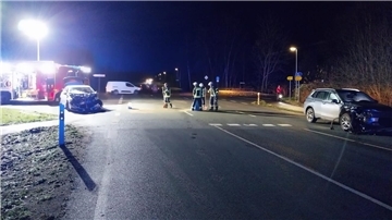 Eine nächtliche Unfallstelle an einer Kreuzung: Zwei stark beschädigte Fahrzeuge stehen auf der Straße, während Feuerwehrleute in Einsatzkleidung die Unfallstelle sichern und Betriebsstoffe aufnehmen. Im Hintergrund ist ein Feuerwehrfahrzeug mit eingeschaltetem Licht zu sehen.