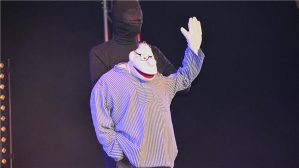 Ein Puppenspieler steht in schwarz gekleidet auf der Bühne und hält eine Stoffpuppe. 