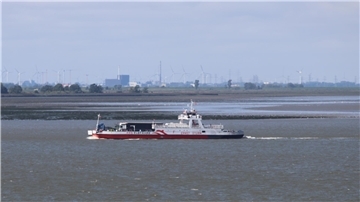 Die Autofähre „Ernst Sturm“ der FRS Elbfähre fährt über die Elbe, im Hintergrund sind bei Ebbe weite Wattflächen sowie Windräder und Industrieanlagen zu sehen.