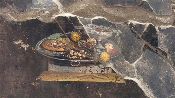 Die Wand eines pompejanischen Hauses mit einem Fresko, das einen Tisch mit Speisen darstellt, darunter auch eine Art Pizza.