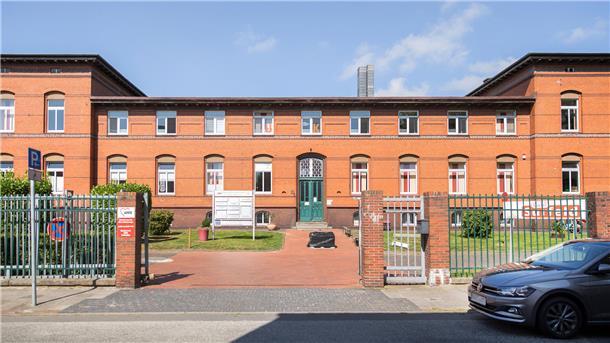 rotes Backsteingebäude mit grüner Tür in einer parkähnlichen Anlage hinter einem Zaun