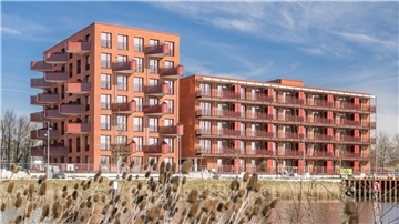 Zwei moderne, rot verklinkerte Wohngebäude mit großzügigen Balkonen am Wasser im Kistner-Carré in Bremerhaven-Lehe. Im Vordergrund Schilfgras, im Hintergrund blauer Himmel mit leichten Wolken.