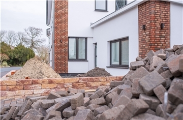 Backsteine liegen aufgehäuft im Vordergrund, dahinter sieht man die Baustelle einer Terrasse und ein frisch saniertes, weiß verputztes Haus.