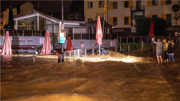 Drei Personen machen Fotos und beobachten die Wassermassen, die sich über eine Straße und ein Restaurant in Euskirchen ausbreiten.