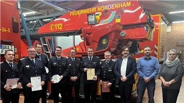 Neun Männer und eine Frau stehen vor Feuerwehrautos in einer Halle