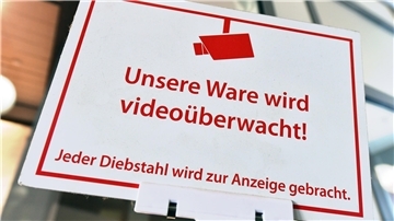Ein Schild mit dieser Aufschrift ist zu sehen: "Unsere Ware wird videoüberwacht! Jeder Diebstahl wird zur Anzeige gebracht." Die Schrift ist rot, das Schild weiß. 