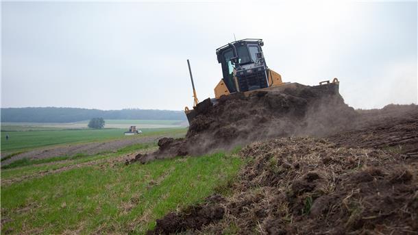 Ein Bagger schält den Mutterboden vom Deich ab. Anschließend wird er mit schwerem Kleiboden erhöht. Am Ende wird der Mutterboden wieder breitgefahren, damit das Gras schneller anwächst.