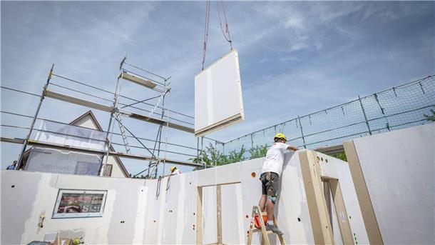 Weniger Fertighäuser gebaut Ein Bauarbeiter steht auf einer Leiter und arbeitet im Rohbau eines Fertighauses, während gerade ein weiteres Wandelement von einem Kran heruntergelassen wird.