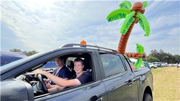 Zwei Männer in einem Auto. Auf der Ladefläche sind zwei Palmen aus Plastik.