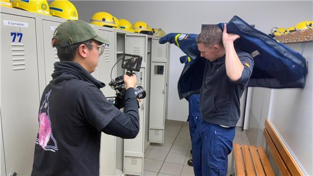 Filmemacher spendet Imagefilm an das THW Bremerhaven  In einer Umkleide eines Hilfswerks zieht sich ein junger Mann eine blaue Einsatzjacke über, während ein Kameramann mit Kappe und schwarzem T-Shirt ihn mit einer professionellen Kamera filmt. Im Hintergrund sind Spinde mit gelben Schutzhelmen zu sehen.