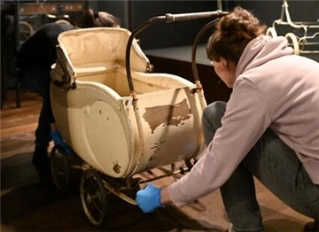 Eine Frau mit blauen Handschuhen schiebt einen alten, beigen Kinderwagen mit abgenutzter Lackierung in eine Museumsausstellung.