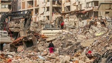 Ein Mann steht in Samandag in der Türkei zwischen Trümmern, während Bagger die Überreste eines zerstörten Gebäudes abtransportieren. Nach dem katastrophalen Beben am 6.2. kommt es in der Region immer wieder zu Nachbeben.