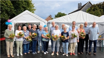 Eine Gruppe von Frauen und Männern steht vor weißen Festzelten, viele von ihnen halten Blumensträuße in den Händen. Im Hintergrund sind bunte Wimpelketten und Luftballons zu sehen.