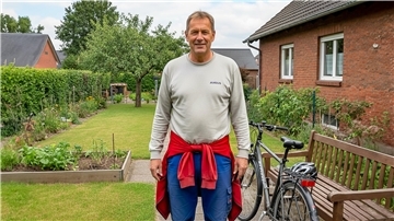 Mit Quiz: Der Durchschnitts-Nordenhamer im Faktencheck Ein Mann steht im Pullover und einer Arbeitshose in einem Garten neben einem Einfamilienhaus. Neben ihm steht ein Fahrrad.
