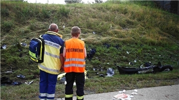 Ein Seelsorger und ein Mitglied der Feuerwehr an einer Unfallstelle auf der Autobahn 9 vor einer Böschung, die mit Trümmerteilen übersäht ist.