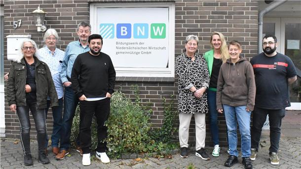 Acht Männer und Frauen stehen vor einer Mauer.