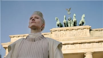 Eine Erlöserfigur im weißen Mantel mit einem Patronengurt um den Bauch - die Künstlerin selbst - schreitet neben einem gescheckten Esel durchs Brandenburger Tor - eine Szene aus Yael Bartanas Filminstallation „Malka Germania“.
