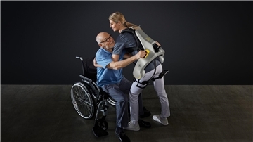 Eine Frau hebt mit Hilfe eines intelligenten Kraftanzugs der Firma German Bionic einen Mann aus einem Rollstuhl.