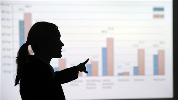 Eine Frau steht in einem Büro und erklärt eine an die Wand projezierte Grafik.